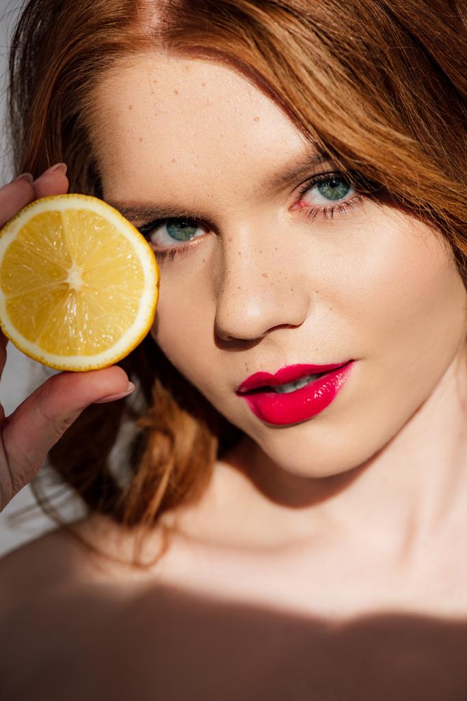 hermosa pelirroja con labios rojos posando con limón cortado y mirando a la cámara
 - Foto, imagen