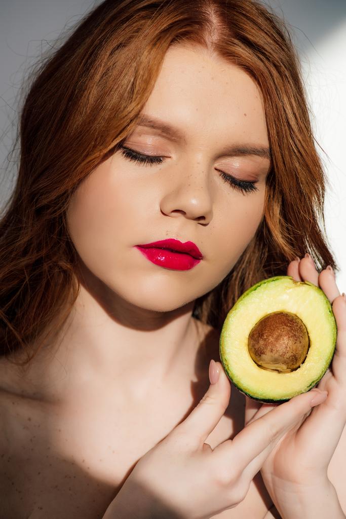 beautiful redhead girl with red lips and eyes closed posing with cut avocado - Photo, Image