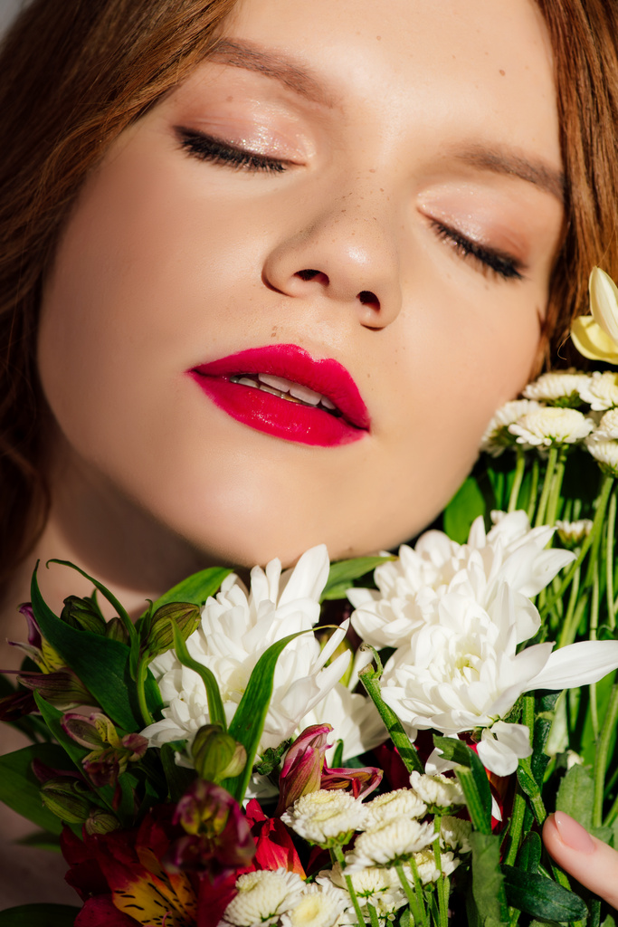 крупный план красивой молодой рыжеволосой женщины с красными губами и закрытыми глазами, позирующей с цветами
 - Фото, изображение