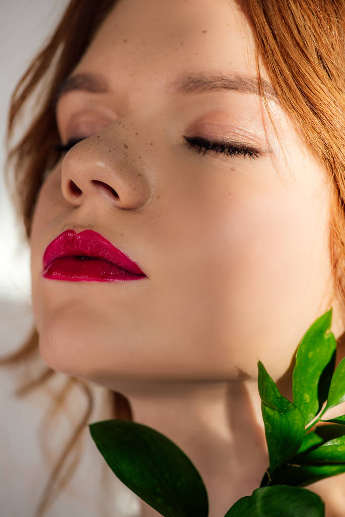 portrait of beautiful young redhead woman with red lips and eyes closed posing with green leaf - Fotografie, Obrázek