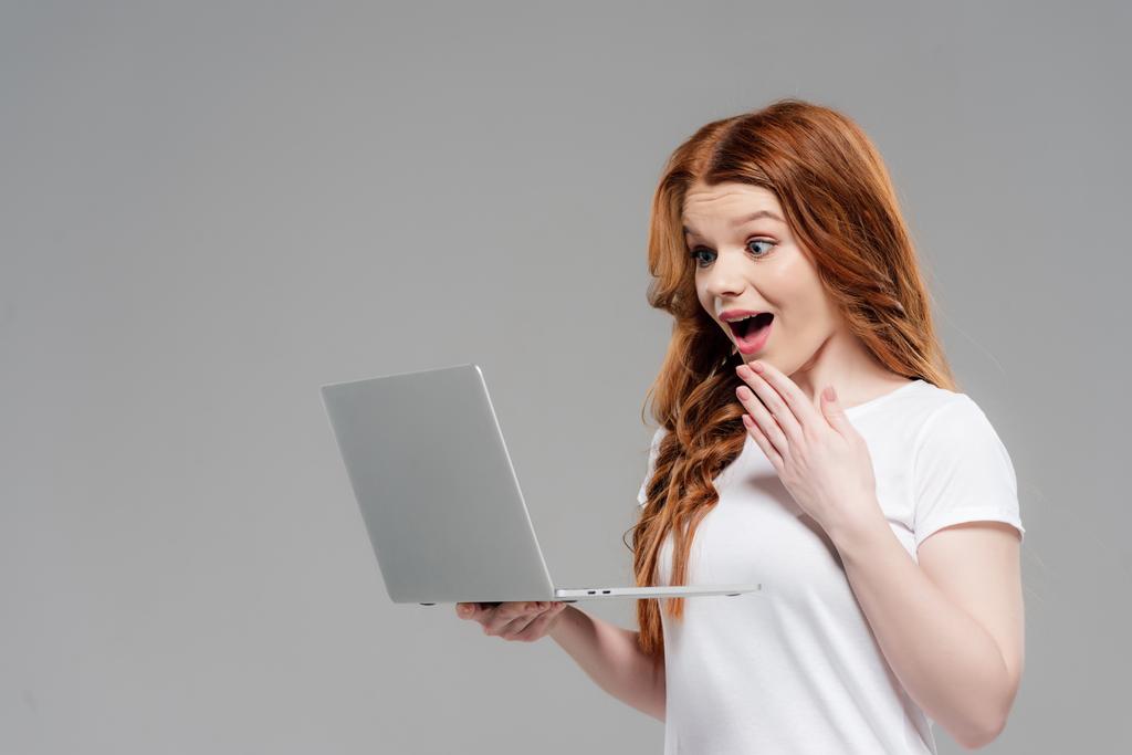 piękne zaskoczony rudowłosy dziewczyna za pomocą laptopa i wskazując z ręki izolowane na szarym - Zdjęcie, obraz