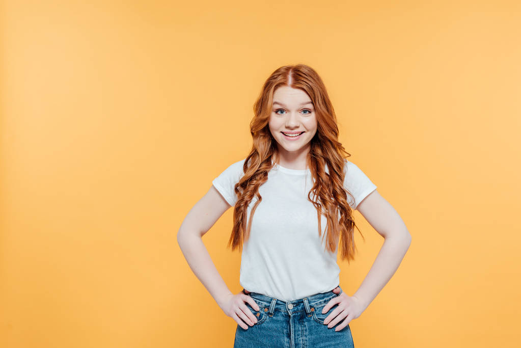 beautiful redhead girl with hands akimbo looking at camera and posing isolated on yellow - Photo, Image