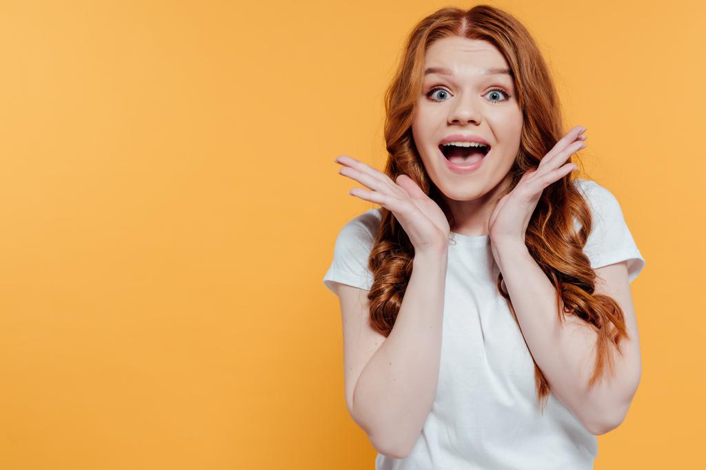 beautiful excited redhead girl looking at camera and gesturing with hands isolated on yellow - Photo, Image