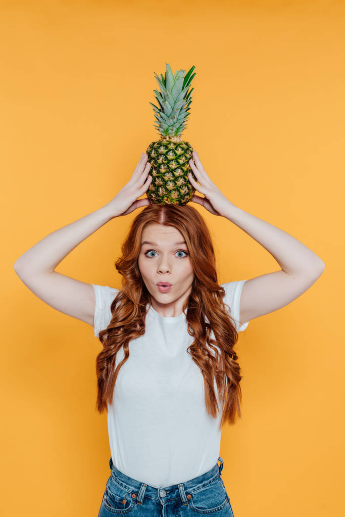 beautiful redhead girl looking at camera, making face expression and posing with pineapple on head isolated on yellow - Photo, Image