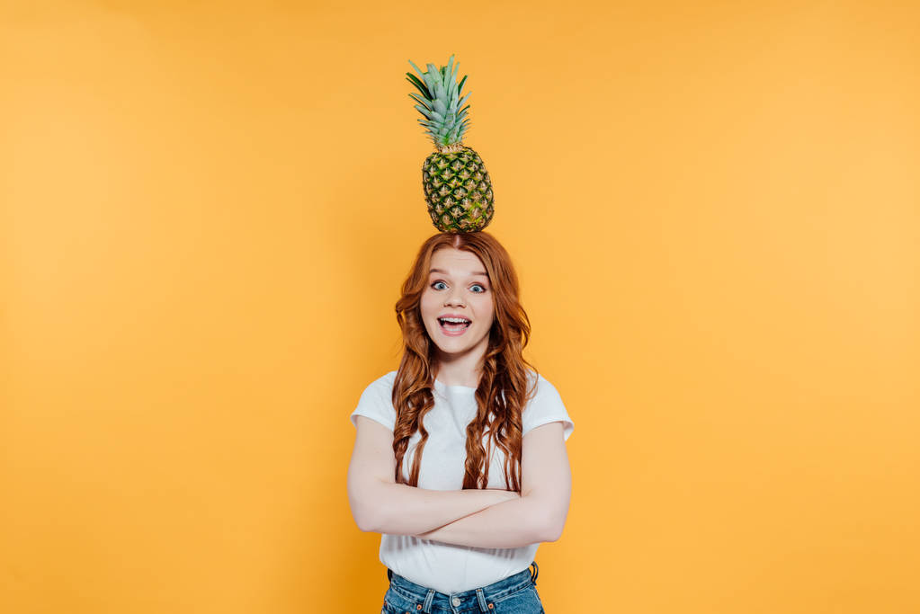 excited redhead girl looking at camera and posing with pineapple on head isolated on yellow - Photo, Image
