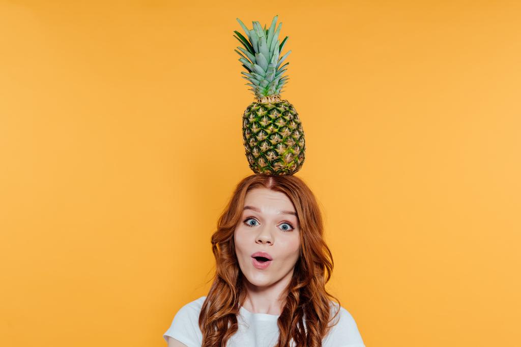 surprised redhead girl looking at camera and posing with pineapple on head isolated on yellow - Photo, Image