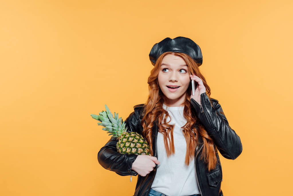 redhead girl in leather jacket holding pineapple and talking on smartphone isolated on yellow with copy space - Photo, Image