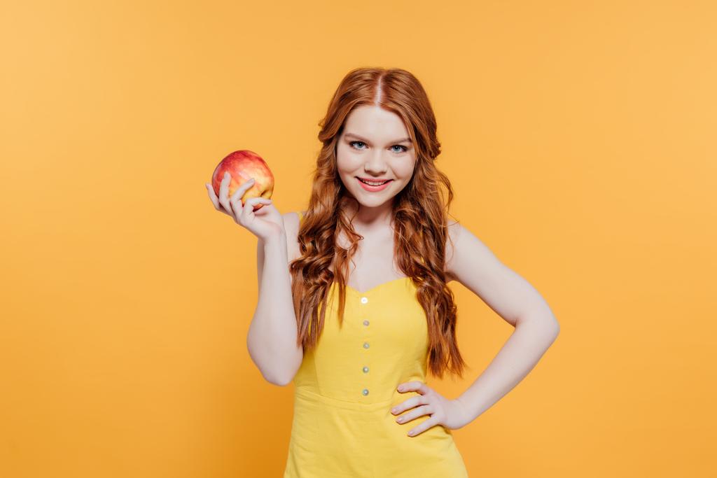 красивая улыбающаяся рыжая девушка с яблоком, смотрящая на камеру, изолированную на желтый
 - Фото, изображение