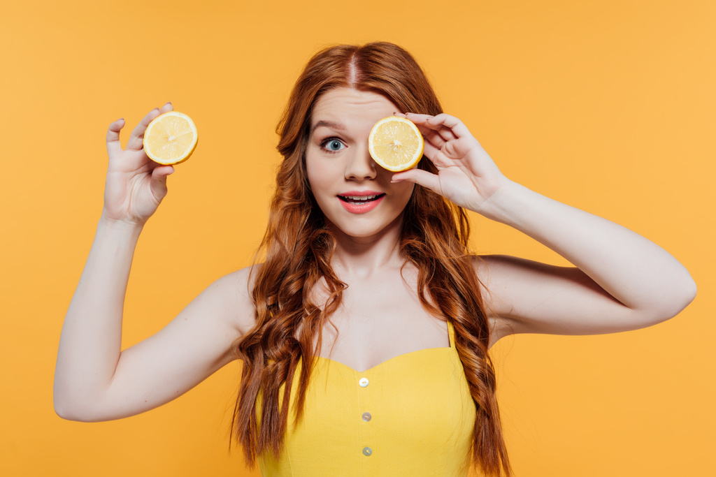 menina ruiva surpresa olhando para a câmera e segurando limão na frente do olho isolado no amarelo
 - Foto, Imagem