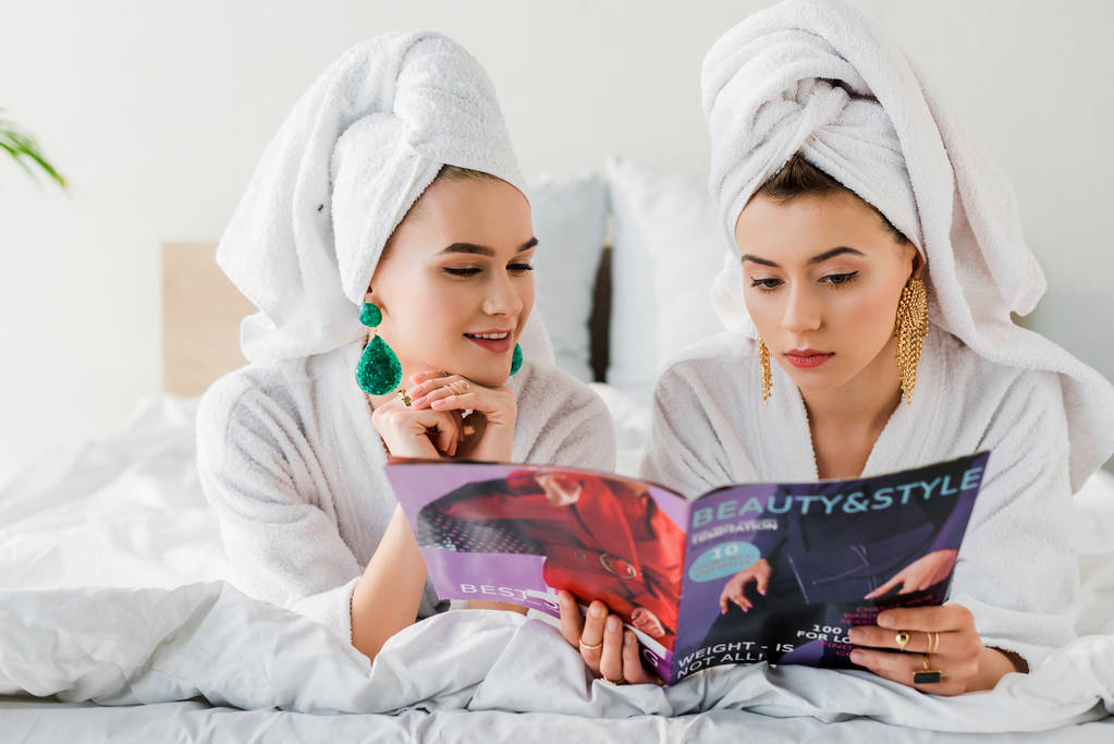mulheres elegantes em roupões de banho, brincos e com toalhas na cabeça ler revista enquanto deitado na cama
 - Foto, Imagem