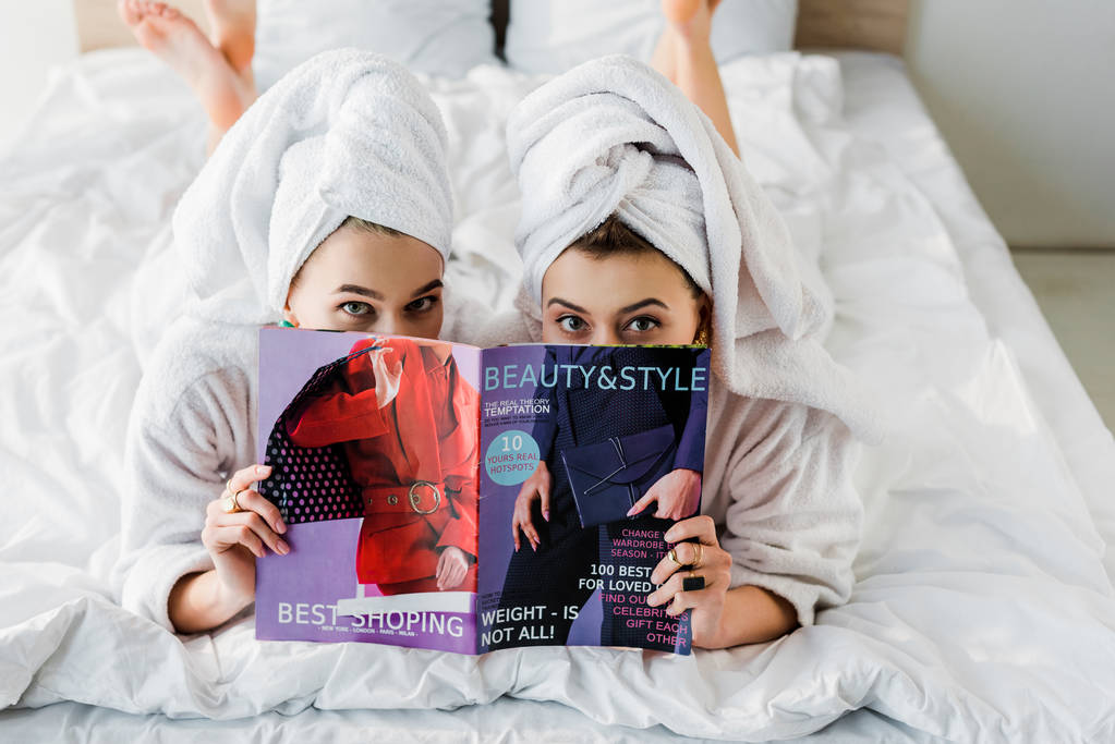 overhead view of barefoot women in bathrobes and with towels on heads hiding behind magazine together while lying in bed - Photo, Image