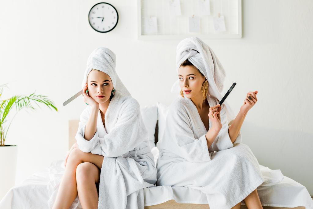 stijlvolle vrouwen in badjassen en sieraden, met handdoeken op de hoofden zittend op bed met nagel bestanden - Foto, afbeelding