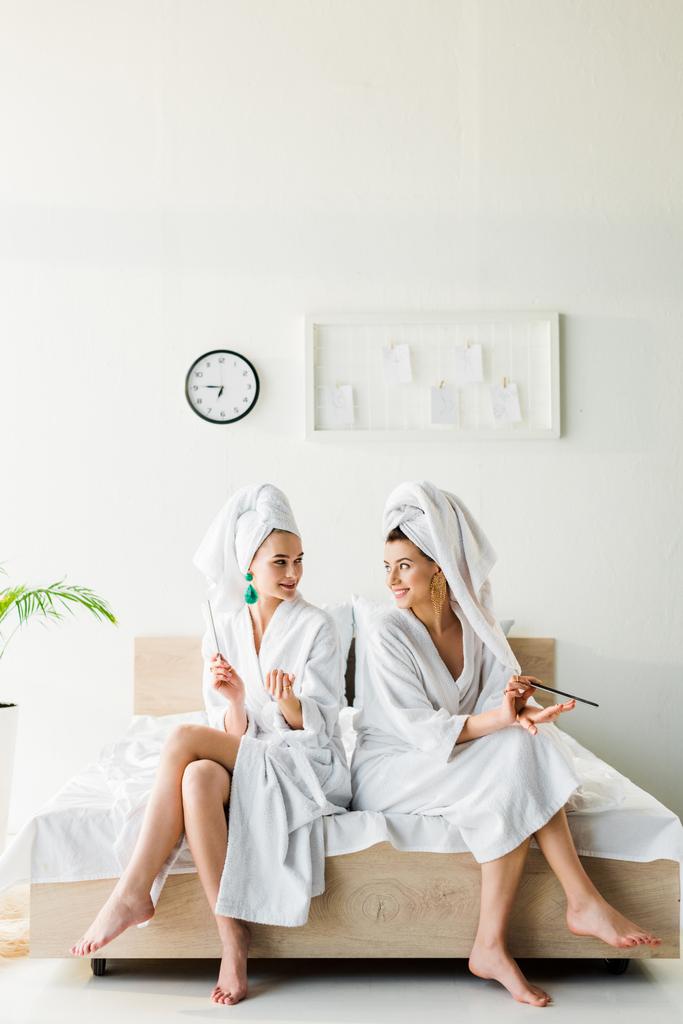 стильные улыбающиеся женщины в халатах и ювелирных украшениях, с полотенцами на головах, сидя на кровати с ногтями
 - Фото, изображение