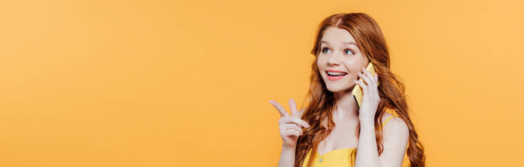 панорамний знімок красивої усміхненої рудої дівчини, що вказує пальцем під час розмови на смартфоні
 - Фото, зображення