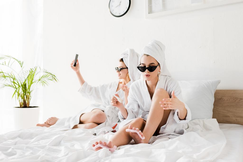 stylish women in bathrobes, sunglasses, towels and jewelry lying in bed, doing pedicure and taking selfie - Photo, Image