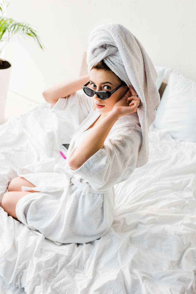 εναέρια θέα της κομψούς γυναίκας με μπουρνούζι και γυαλιά ηλίου, πετσέτα και κοσμήματα που κάθονται στο κρεβάτι - Φωτογραφία, εικόνα