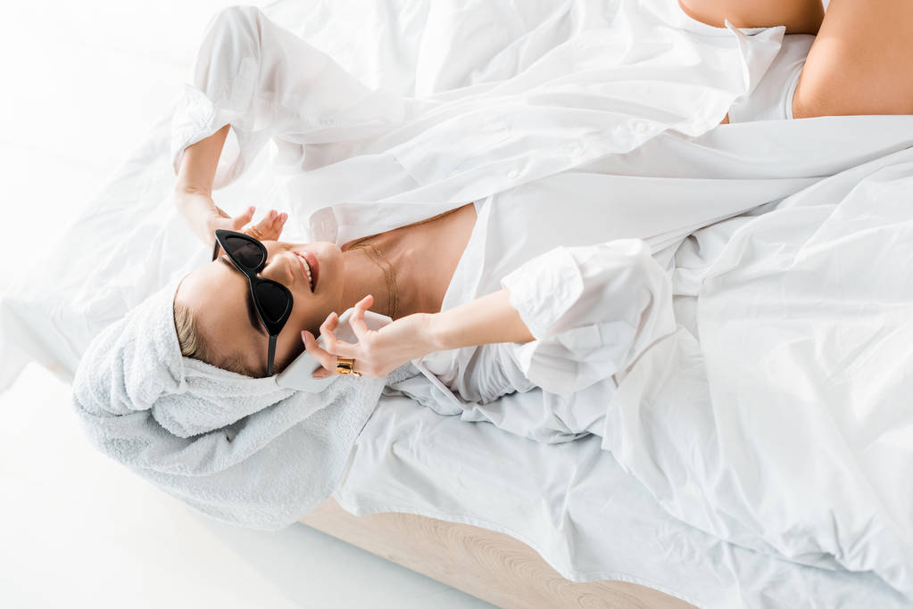 молодая улыбающаяся женщина в рубашке, солнцезащитных очках, ювелирных украшениях и с полотенцем на голове разговаривает на смартфоне в постели
 - Фото, изображение