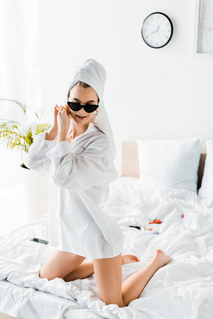 jeune femme élégante en chemise, lunettes de soleil, bijoux et avec serviette sur la tête mettre une boucle d'oreille dans le lit
 - Photo, image
