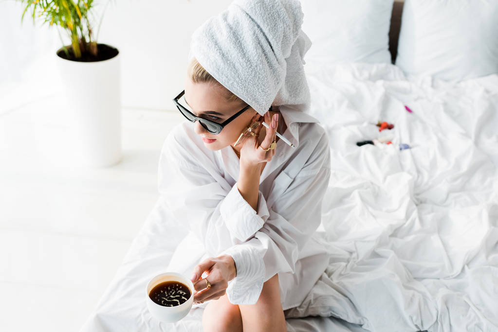 fiatal elegáns nő ékszerek és napszemüveg törölközővel a fején és csésze kávé dohányzás cigaretta, miközben ül az ágyon - Fotó, kép
