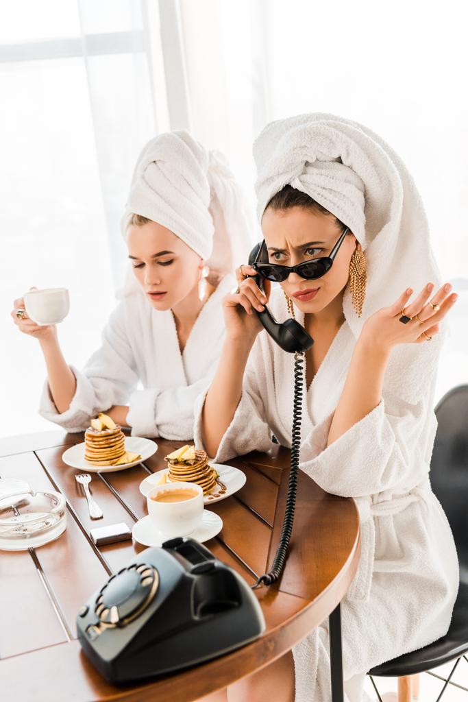 κομψή δυσαρεστημένη γυναίκα σε μπουρνούζι, γυαλιά ηλίου και κοσμήματα με πετσέτα στο κεφάλι μιλάει σε ρετρό τηλέφωνο, ενώ έχει πρωινό με φίλο - Φωτογραφία, εικόνα