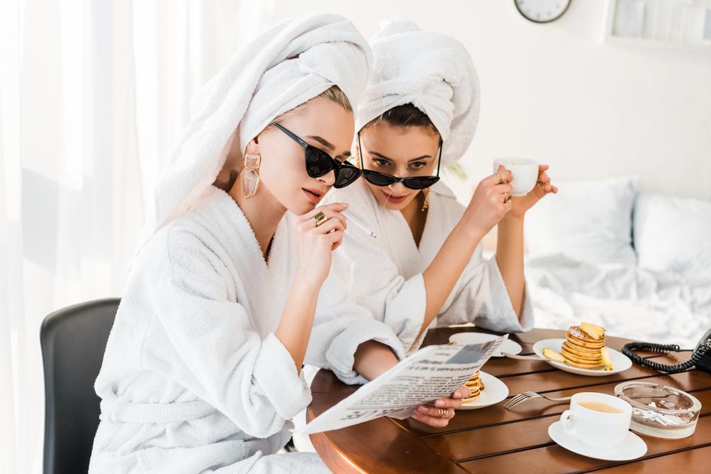femmes élégantes en peignoirs, lunettes de soleil et bijoux avec des serviettes sur les têtes fumer et lire le journal tout en prenant le petit déjeuner
  - Photo, image