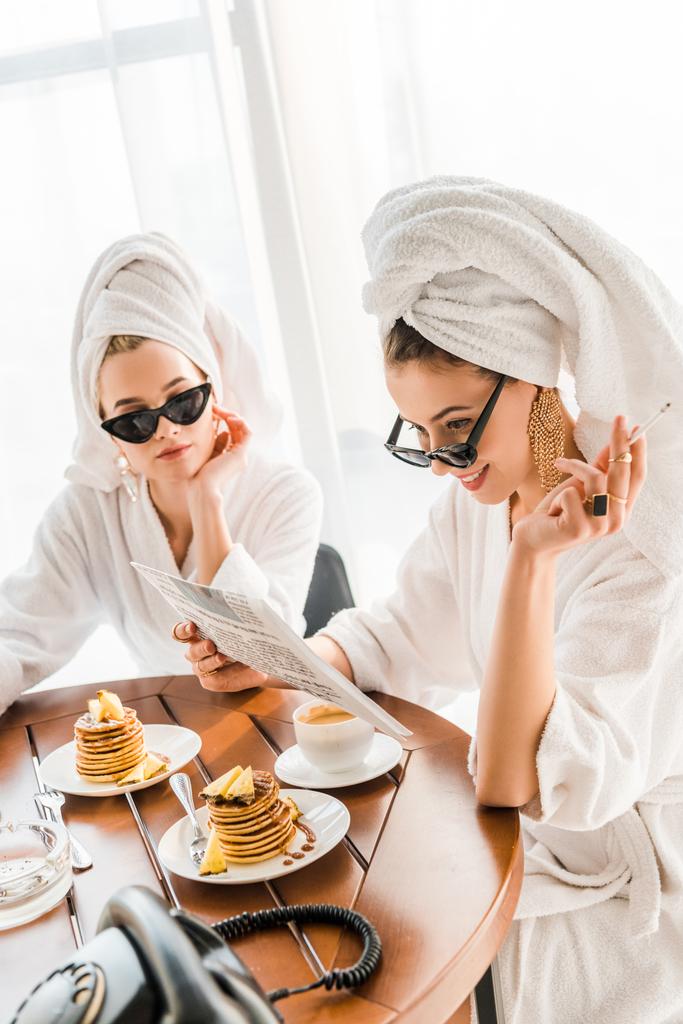 стильная улыбающаяся женщина в халате, солнцезащитных очках и украшениях с полотенцем на голове, курящей сигарету и читающей газету с другом по утрам
 - Фото, изображение