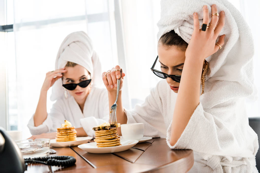 stijlvolle vermoeide vrouwen in badjassen, zonnebrillen en sieraden met handdoeken op hoofden eten pannenkoeken voor het ontbijt - Foto, afbeelding