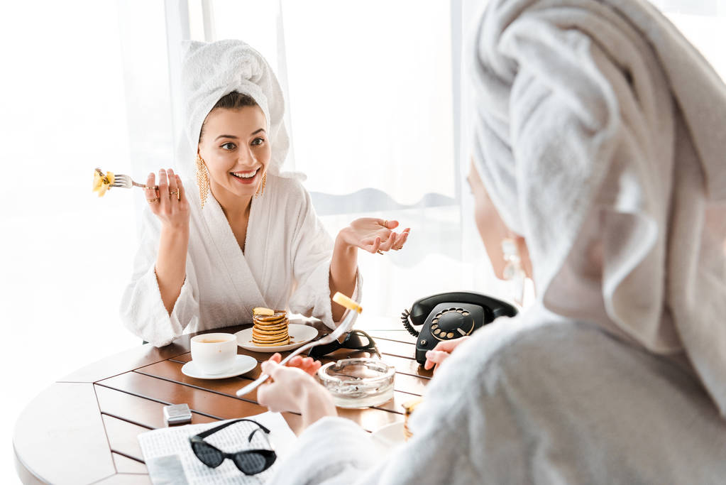 стильные улыбающиеся женщины в халатах и украшениях с полотенцами на головах разговаривают во время завтрака
 - Фото, изображение