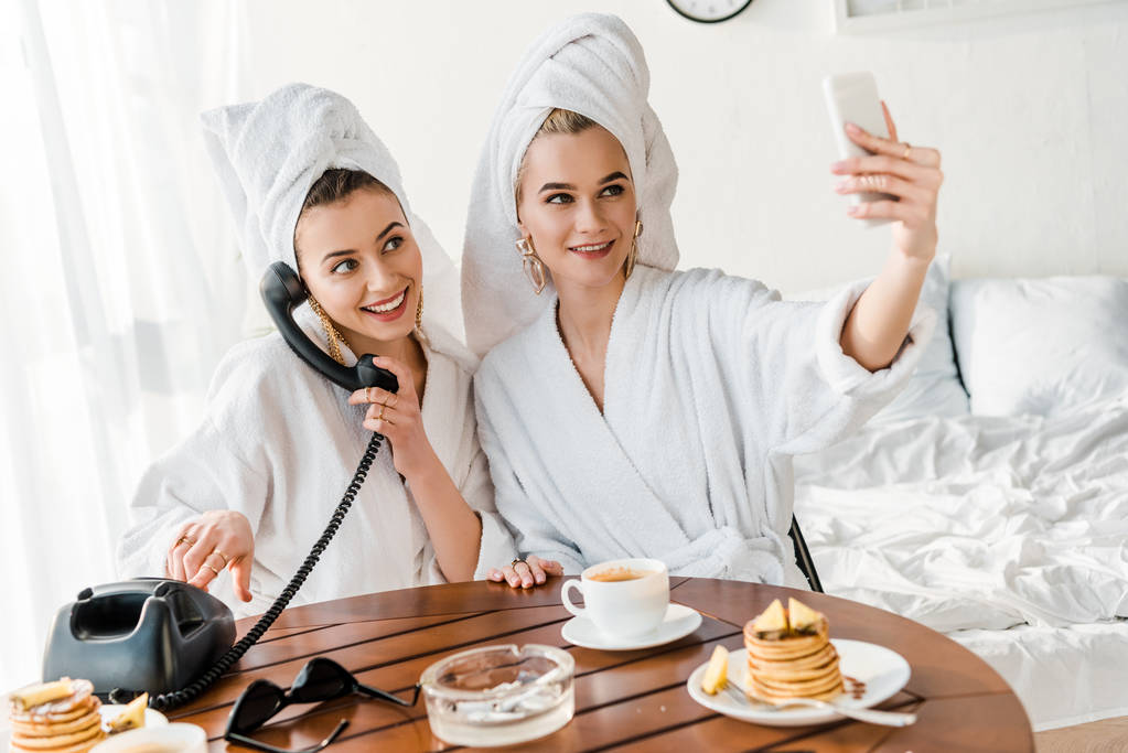 tyylikkäät naiset kylpytakit ja korut pyyhkeillä päissä puhuvat retro-puhelimessa ja hymyilevät selfien ottamisen aikana
 - Valokuva, kuva