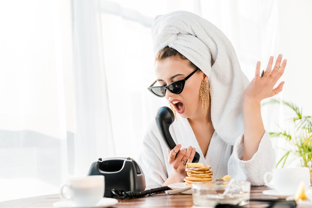 irritated stylish woman in bathrobe, sunglasses and jewelry with towel on head screaming and gesturing while talking on retro telephone  - Photo, Image