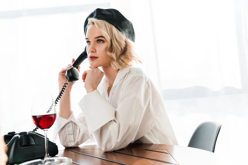 elegante blonde Frau in schwarzer Baskenmütze und weißem Hemd sitzt am Tisch und spricht mit einem Retro-Telefon neben Glas mit Rotwein auf dem Tisch - Foto, Bild