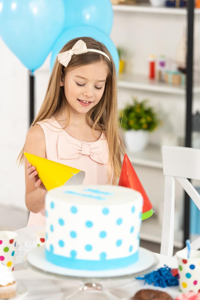 adorable kid holding party cap near table with cake during birthday celebration - Photo, Image