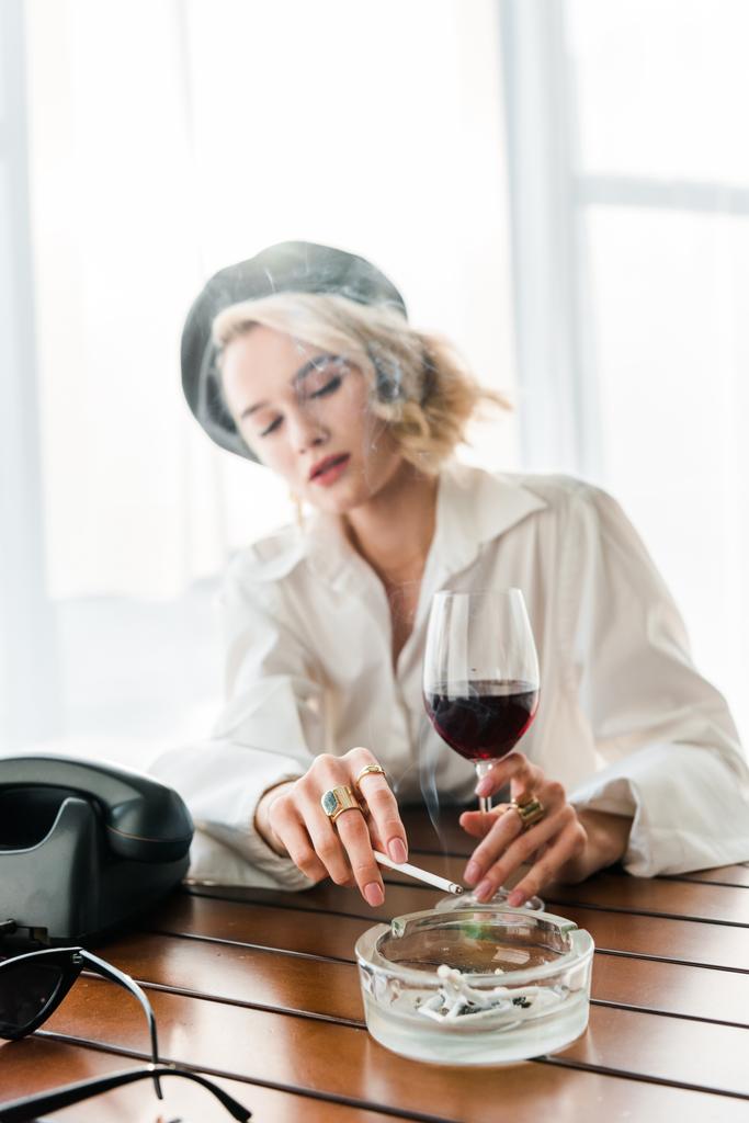 Selektiver Fokus der eleganten blonden Frau in schwarzer Baskenmütze, Zigarette rauchend und Glas mit Rotwein in der Hand  - Foto, Bild