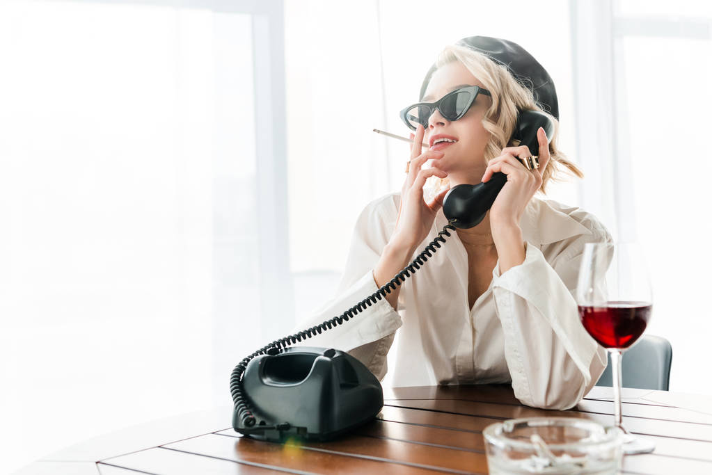 élégante femme blonde souriante en béret noir et lunettes de soleil fumant cigarette tout en parlant sur téléphone rétro près d'un verre de vin rouge
 - Photo, image