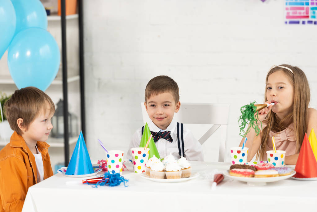 Kinder sitzen bei Geburtstagsfeier mit Cupcakes und Partyhörnern am Tisch - Foto, Bild