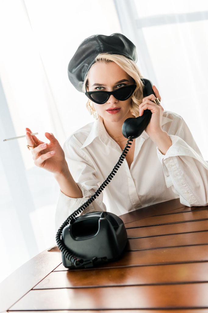 élégante femme blonde en béret noir et lunettes de soleil fumer cigarette tout en parlant sur téléphone rétro
  - Photo, image