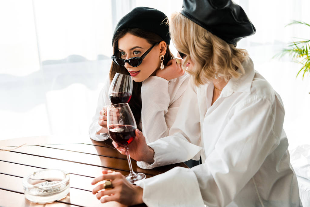 Elegancka brunetka i blond kobiety w czarnych Beretów i okulary do picia czerwone wino na drewnianym stole - Zdjęcie, obraz