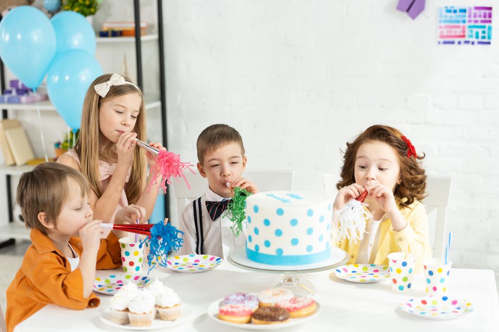 kinderen zitten aan tafel met cake en blazen partij hoorns tijdens de verjaardag viering - Foto, afbeelding