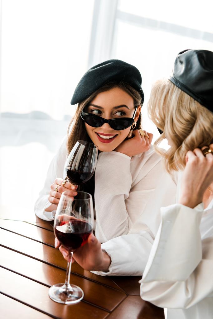 mulher morena sorridente elegante em boina preta e óculos de sol bebendo vinho tinto na mesa de madeira com amigo loiro
 - Foto, Imagem