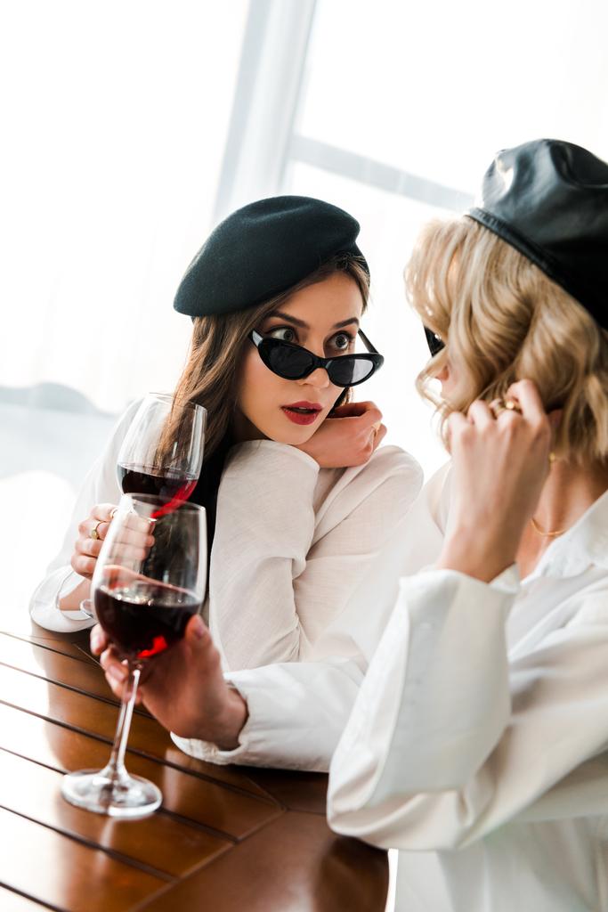 elegante geschokt brunette vrouw in zwarte baret en zonnebril drinken rode wijn op houten tafel met blonde vriend - Foto, afbeelding