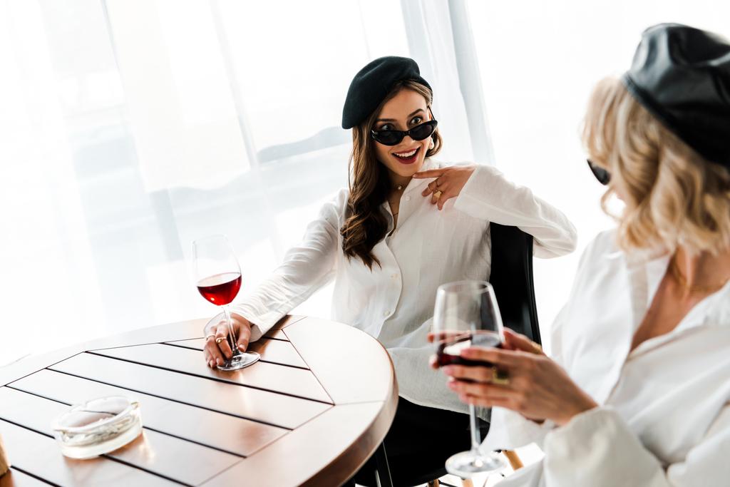 elegante mujer morena sonriente y sorprendida en boina negra y gafas de sol bebiendo vino tinto en mesa de madera con amiga rubia
 - Foto, imagen