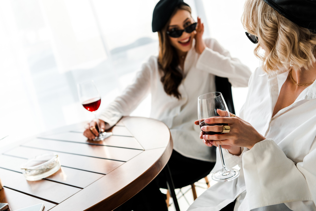 selective focus of blonde woman drinking wine with elegant smiling and surprised brunette friend in black beret and sunglasses  - Photo, Image