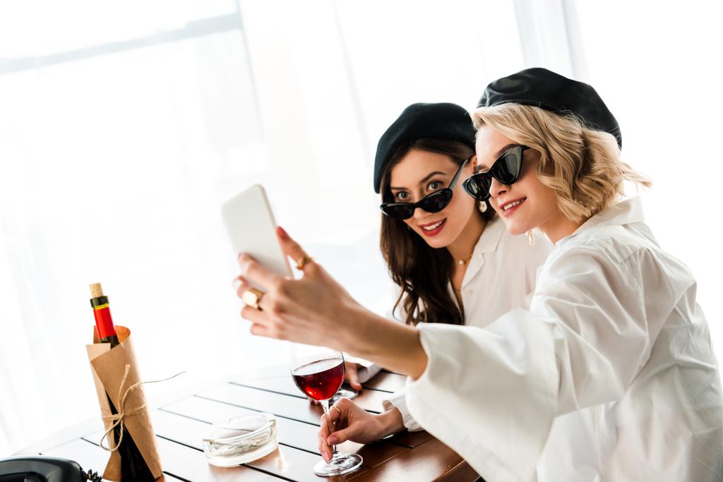 κομψό χαμογελαστή μελαχρινή και ξανθιές γυναίκες σε μαύρους μπερέδες και γυαλιά ηλίου πίνοντας κόκκινο κρασί και παίρνοντας selfie - Φωτογραφία, εικόνα