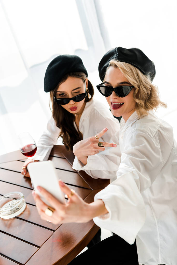 elegantní bruneta a blonďaté ženy v černých baretech a slunečních brýlích, které popíjejí červené víno a berou si na sebe slast, a ukazují vítězné znamení - Fotografie, Obrázek