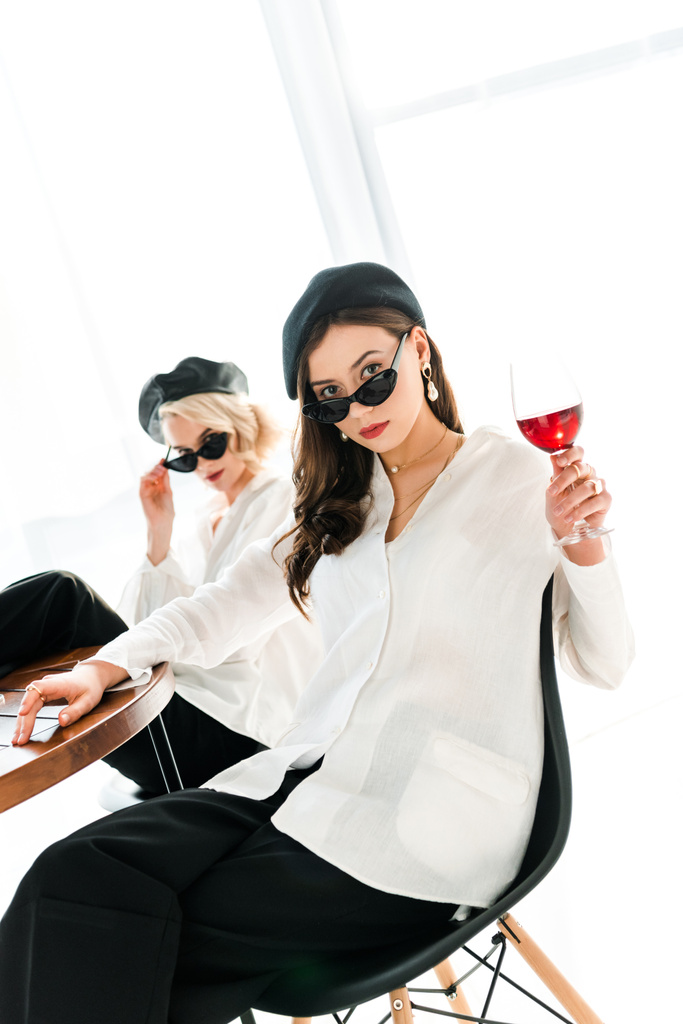selektywne skupienie eleganckiej kobiety brunetka w czarnym Beret i okulary przeciwsłoneczne z kieliszek czerwonego wina siedzi w pobliżu blond przyjaciela - Zdjęcie, obraz