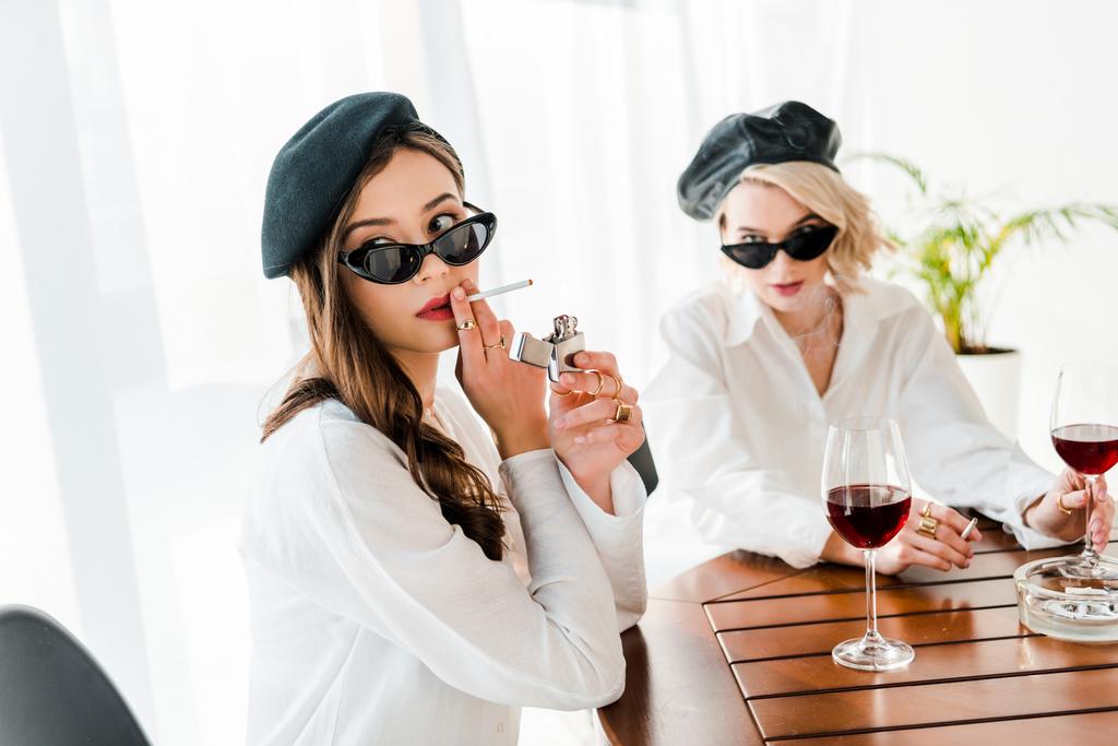 selective focus of elegant woman in black beret and sunglasses lighting up cigarette near friend  - Photo, Image