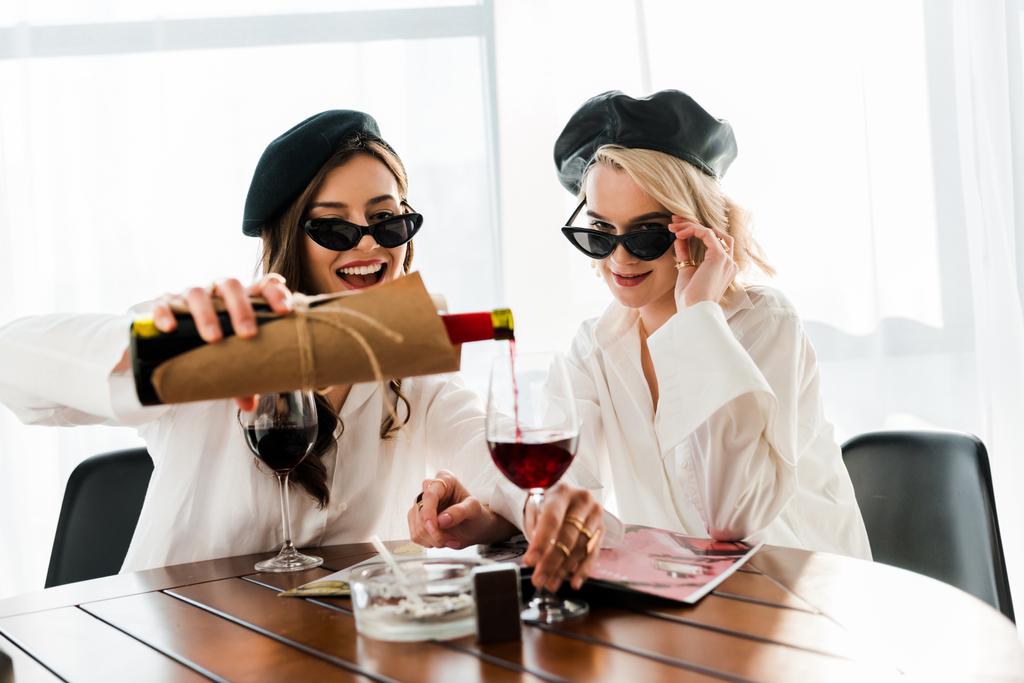 esmer ve sarışın mutlu kadınlar siyah bere ve güneş gözlüğü kırmızı şarap ve okuma dergisi dökme  - Fotoğraf, Görsel
