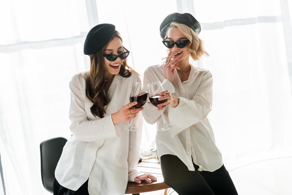 Elegancka szczęśliwa brunetka i blond kobiety w czarnych Beretów i okulary przeciwsłoneczne śmiech, palenie i picie czerwonego wina - Zdjęcie, obraz