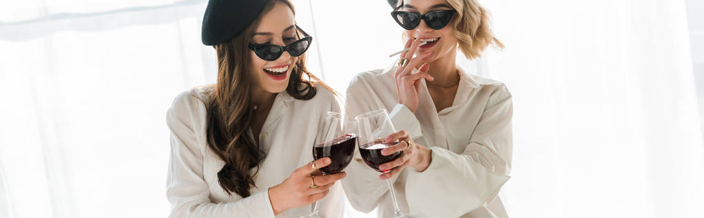 panoramische shot van elegante gelukkige brunette en blonde vrouwen in zwarte baretten en zonnebrillen lachen, roken en drinken van rode wijn - Foto, afbeelding