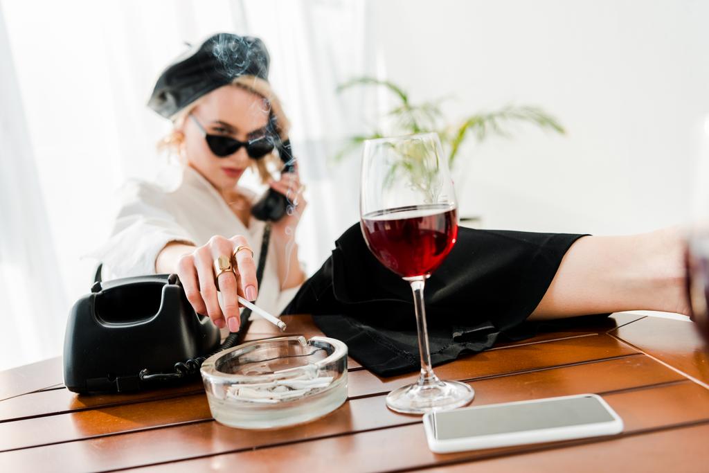siyah bere ve güneş gözlüğü sigara ciddi zarif sarışın kadın seçici odak ve kırmızı şarap ile cam yakınında retro telefon konuşan - Fotoğraf, Görsel